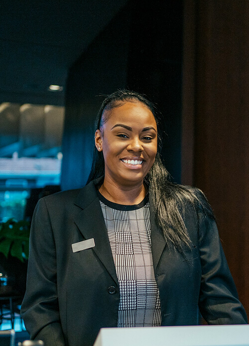 Person in a black blazer smiling at the camera, standing indoors in front of a dark background with soft lighting.