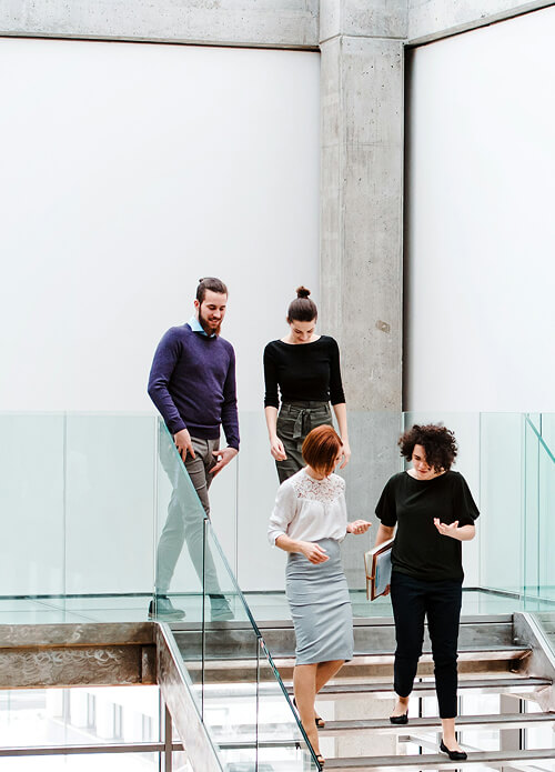Four people are walking down a modern, glass-paneled staircase. Two women are in front, engaged in conversation, followed by two men. The background consists of minimalist, concrete walls.