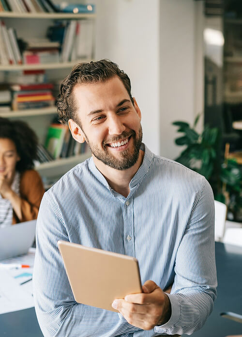 A man in a striped shirt holds a tablet and smiles in an office with bookshelves and plants. A person works on a laptop in the background.