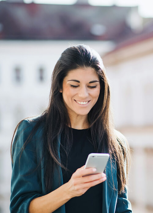 Woman with long hair wearing a dark blazer, smiling while looking at her smartphone outdoors on a sunny day.