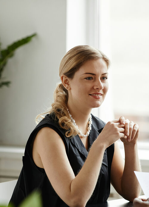 A person with blonde hair in a ponytail, wearing a black sleeveless top and a pearl necklace, sits at a table near a window with a subtle smile.