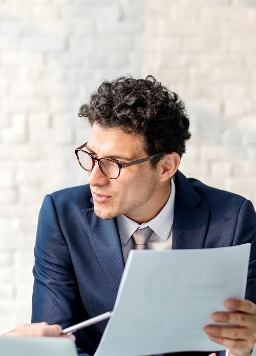A man in glasses and a suit holds a document, seated at a table against a light brick wall backdrop.