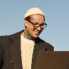 Person wearing glasses, a beige beanie, cable-knit sweater, and dark coat, smiles while looking at a laptop screen against a clear blue sky.