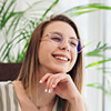 A person with glasses smiling, sitting indoors with plants in the background.