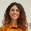 Smiling person with curly hair and glasses wearing an orange shirt against a plain background.