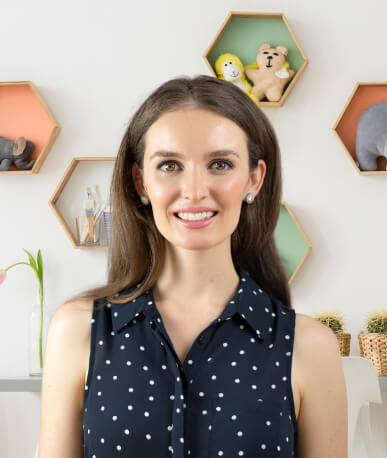A woman with long brown hair and a sleeveless black polka dot top smiles at the camera; hexagonal shelves with toys and decor are on the wall behind her.