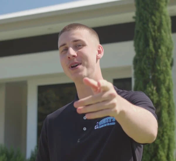 A man in a black polo shirt is standing outdoors in front of a building, pointing towards the camera and smiling.