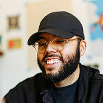 A bearded person wearing glasses and a black cap smiles indoors, with blurred artwork visible in the background.