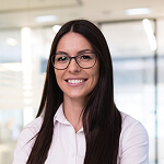 Woman with long dark hair and glasses, wearing a white blouse, standing indoors in an office environment, smiling at the camera.