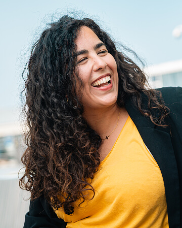 A woman with long curly hair and a yellow top smiles and laughs outdoors, wearing a black jacket.