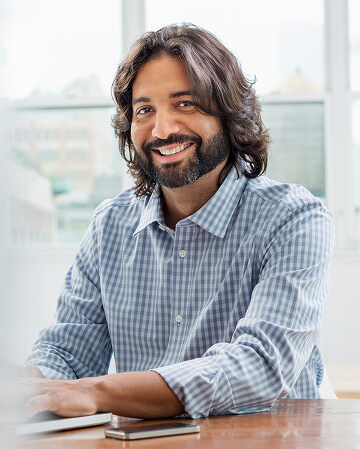A man with shoulder-length hair and a beard, wearing a checked shirt, sits at a desk and smiles at the camera with a smartphone in front of him.