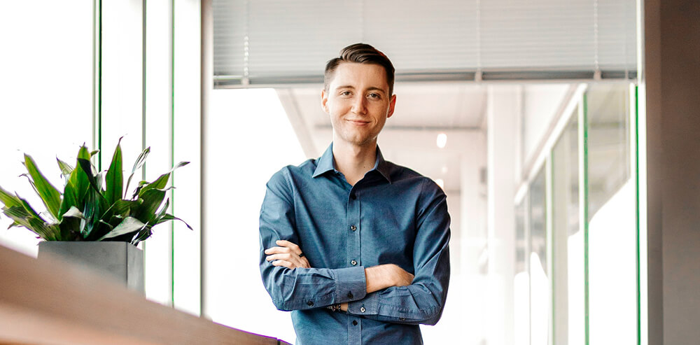 A man in a blue shirt stands indoors with his arms crossed, smiling, next to a potted plant and large windows.