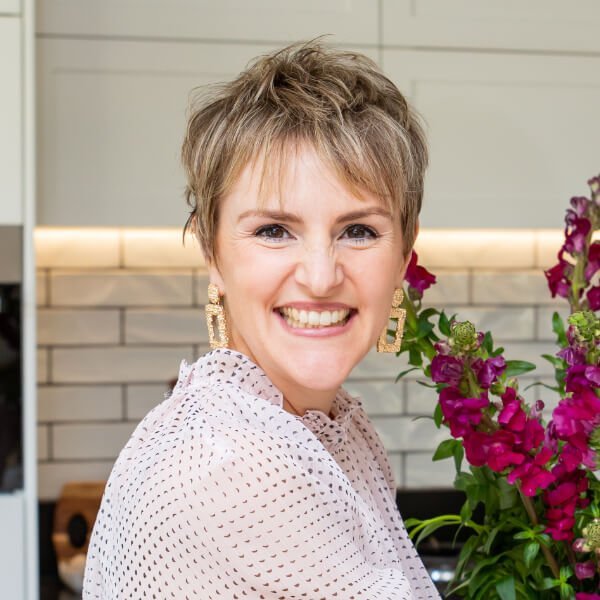 A smiling woman with short hair and large earrings stands indoors next to a vase of vibrant purple flowers.