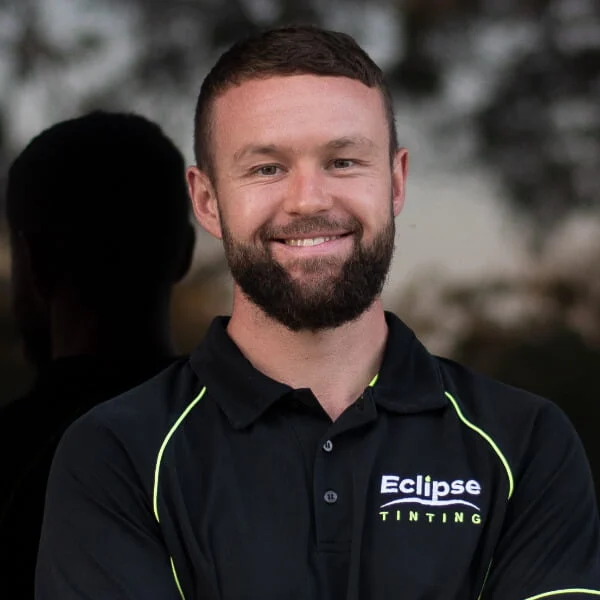 A man with a beard and short hair stands outdoors, smiling with arms crossed, wearing a black "Eclipse Tinting" polo shirt.